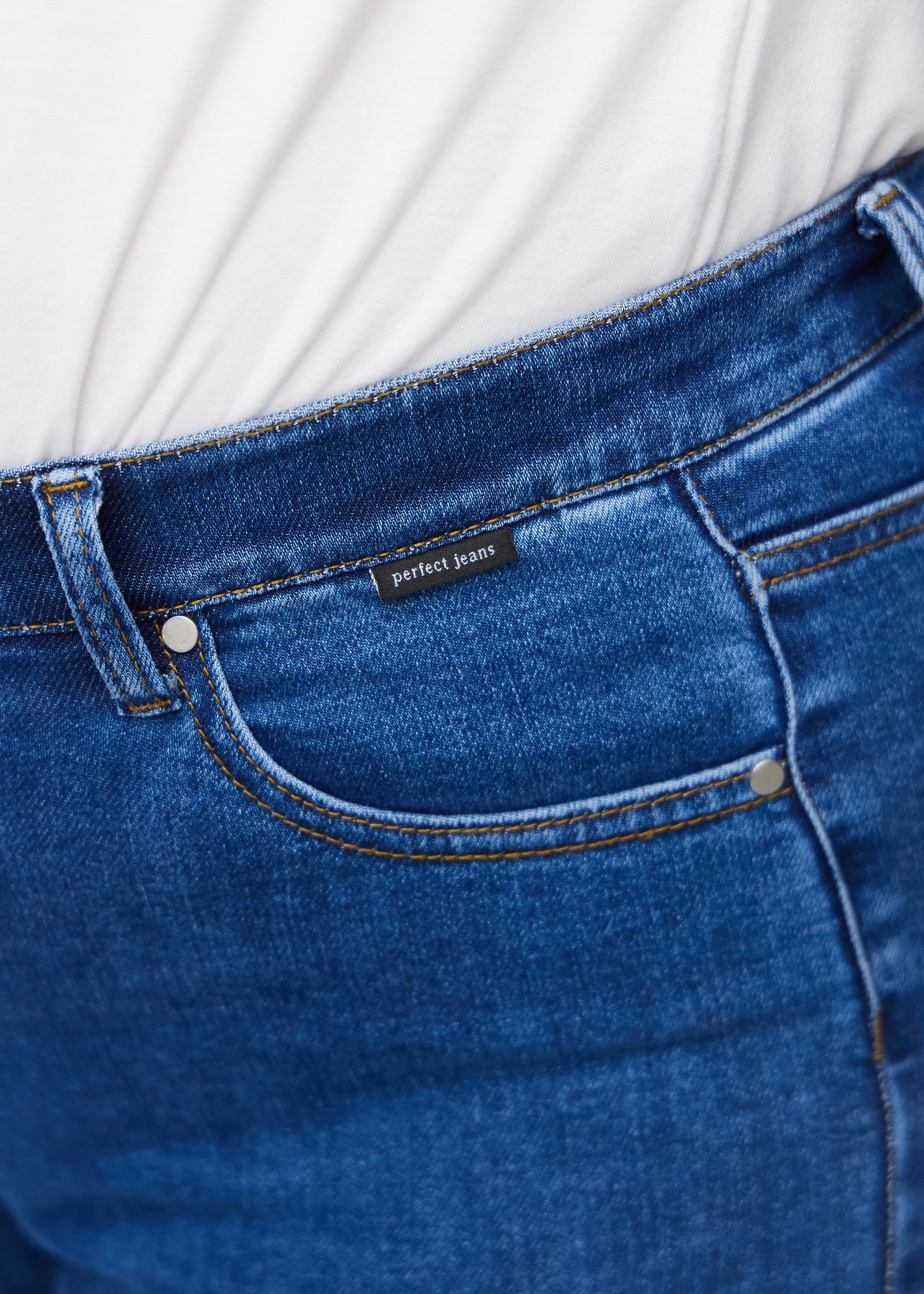 Close-up of dark blue denim with the Perfect Jeans logo label at the edge of the front pocket on a plus-size model.