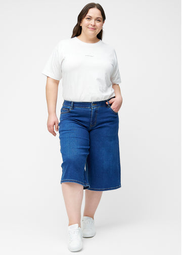 Fuldt billede af plus-size model i løse, mørkeblå denim capris i farven Oceans, der går til lige under knæet.