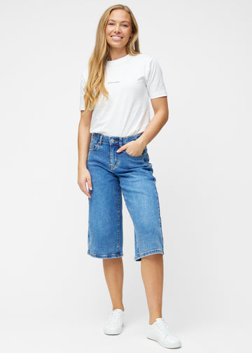 Fuldt billede af model i løse, klassisk blå denim capris i farven Rivers, der går til lige under knæet.