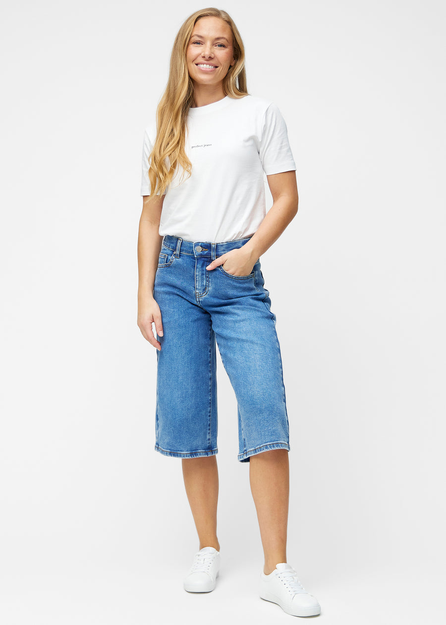 Fuldt billede af model i løse, klassisk blå denim capris i farven Rivers, der går til lige under knæet.