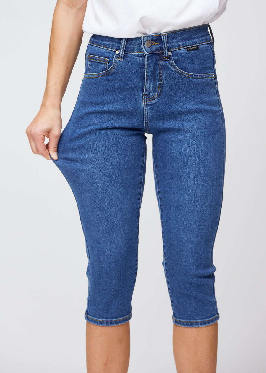 Slim capris for women in dark blue denim, color Oceans. A model pulls the fabric at the thigh to demonstrate stretch and flexibility.