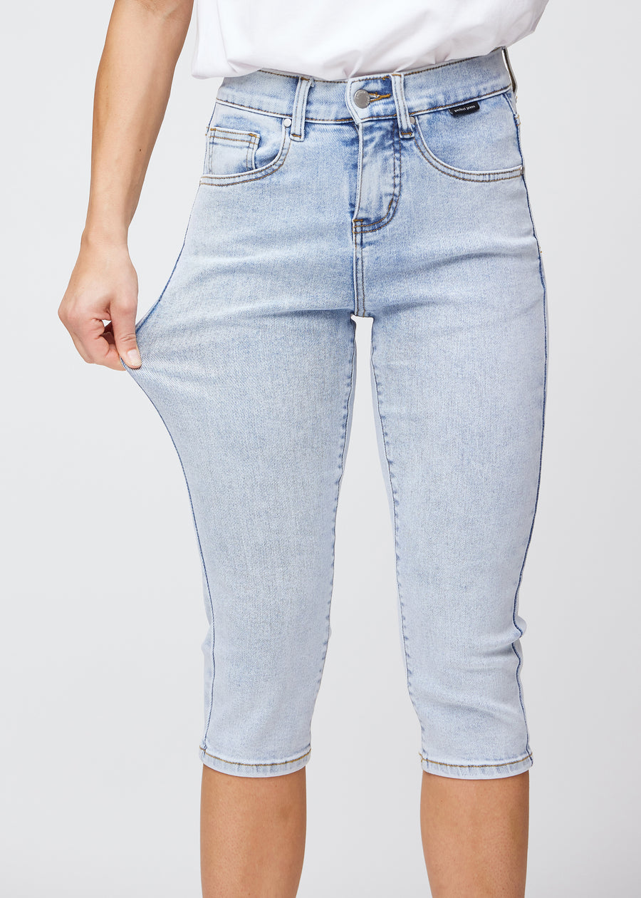 Slim capris for women in light blue denim, color Waves. A model pulls the fabric at the thigh to demonstrate stretch and flexibility.