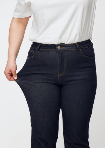 A plus-size model stretches the deep dark blue denim jeans for women in the color Midnights at the thigh to show the material&